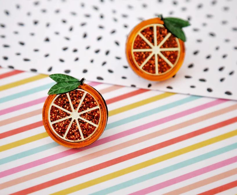 A pair of "Orange You Nice" hoop earrings by Blue Dog Posters, featuring handmade orange slice shapes with glittery interiors and green leaf accents, are placed against a backdrop of polka-dot and rainbow-striped patterns. The earrings include surgical steel findings for a delicate touch.