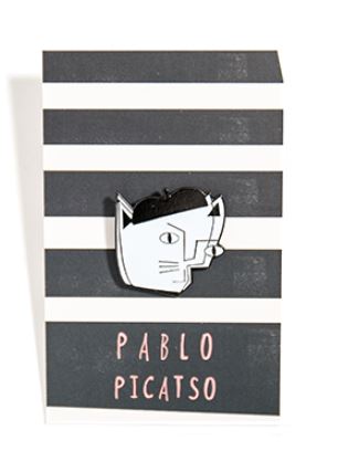 A stylish enamel pin featuring a minimalist face in black and white is attached to a striped card background. Below the pin, the text reads "Pablo Picatso Enamel Pin" in pink letters, with the face resembling Picasso's abstract art style. Designed by Nia Gould, this piece is a quirky nod to Pablo Picasso's genius. Available from Blue Dog Posters.