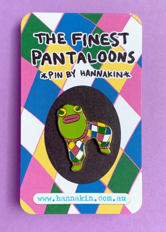 The Finest Pantaloons Enamel Pin by Blue Dog Posters is a vibrant hard enamel pin showcasing a green frog in multicolored diamond-patterned pants. The pin comes on a card with a matching background and gold metal finish. The card features the text "The Finest Pantaloons" and "Pin by Hannakin." For more information, visit hannakin.com.au.