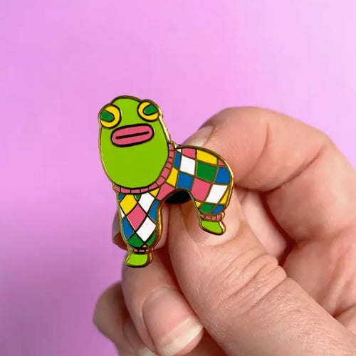 A hand holds "The Finest Pantaloons Enamel Pin" by Blue Dog Posters, shaped like a cartoon frog wearing a brightly patterned harlequin suit with diamond shapes in various colors against a pink background. The frog's body is green, it has a pink mouth, and the pin features a stunning gold metal finish.