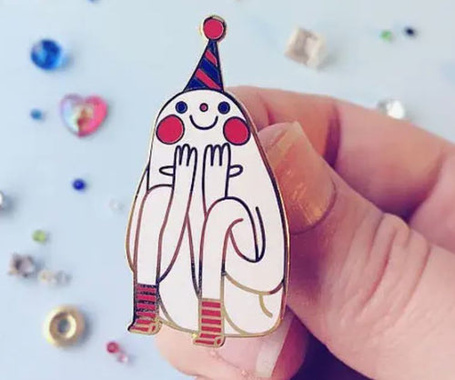 A hand is holding a charming Blue Dog Posters Party Pin Enamel Pin, showcasing an adorable white character with rosy cheeks, a smile, and a conical striped party hat. The hard enamel pin features the character with hands on its cheeks and wearing striped socks. In the background, colorful beads and small objects add charm to the scene.