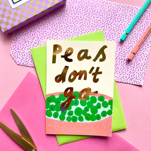 The Peas Don't Go Greeting Card by Blue Dog Posters features gold lettering above a bowl of illustrated green peas, with pink and green envelopes, pencils, scissors, and a patterned box arranged on a pink surface.
