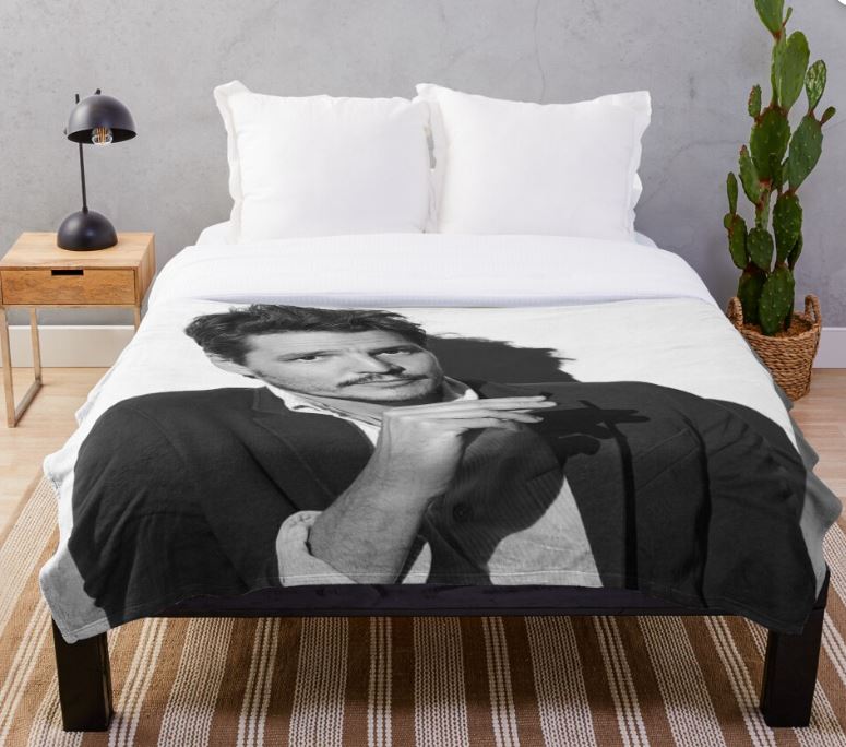 A neatly made bed draped with the Pedro Pascal Throw Blanket from Blue Dog Posters, featuring a black-and-white image of a man with a mustache holding his finger to his lips in a shushing gesture. The machine-washable, 100% polyester bedroom accessory is complemented by a side table, lamp, and potted cactus against a gray wall.