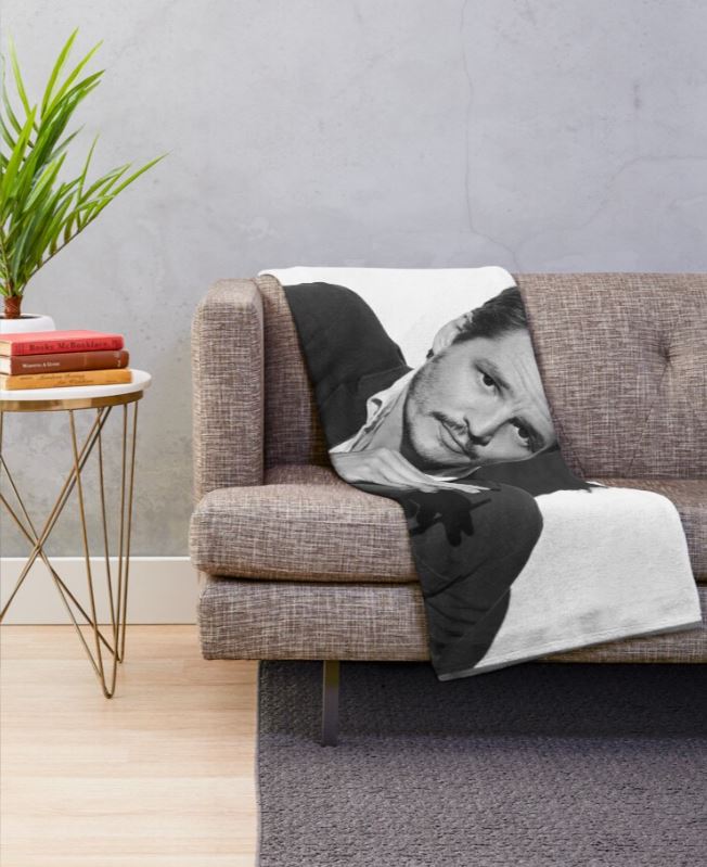 A cozy, gray couch adorned with a soft, fluffy Blue Dog Posters' Pedro Pascal Throw Blanket features an image of a man lying on it. The 100% polyester blanket is machine washable for convenience. Beside the couch stands a small round table with an array of books and a potted plant, all set against a white wall with wooden flooring.