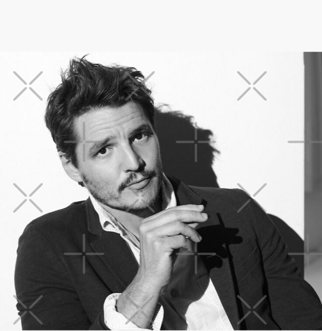A black-and-white photo features a man with a slight beard and mustache sitting against a plain background. He is wearing a blazer over a shirt, holding an object near his face, and draped in the luxurious Pedro Pascal Throw Blanket by Blue Dog Posters. His expression is serious, and his shadow is visible on the wall behind him.
