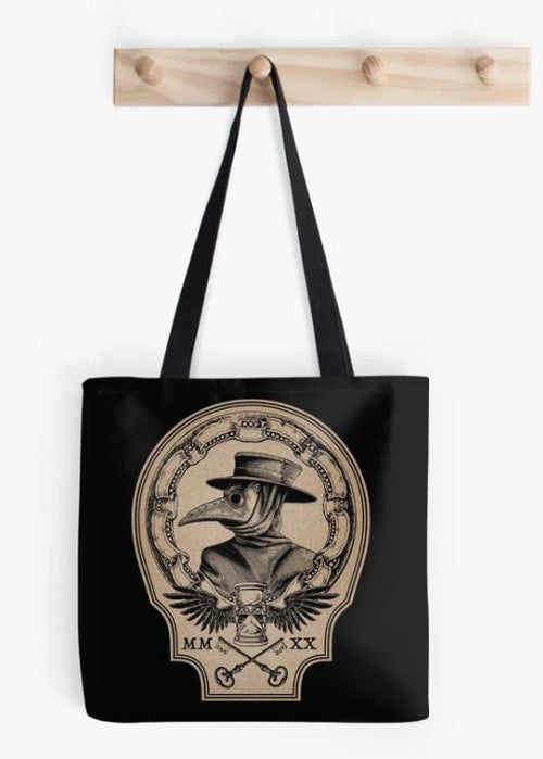 The Vintage Plague Doctor Tote Bag by Blue Dog Posters, made from quality blended fabric, hangs from a wooden peg rack. The black bag features an illustration of a figure wearing a plague doctor mask and wide-brimmed hat, framed by an ornate oval border with a chain design. Beneath the sublimation printed illustration are the Roman numerals "MM" on the left and "XX" on the right.