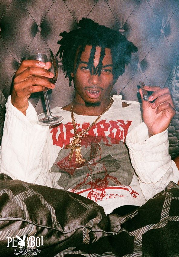 A person with dreadlocks, holding a wine glass and smoking, poses in a distressed shirt decorated with graphics and necklaces. They sit against a tufted headboard, radiating cool confidence. The corner of the bed showcases the "PlayBoi Carti" text logo from MUS 349, evoking an image akin to a rap artist poster from an eclectic music collection.