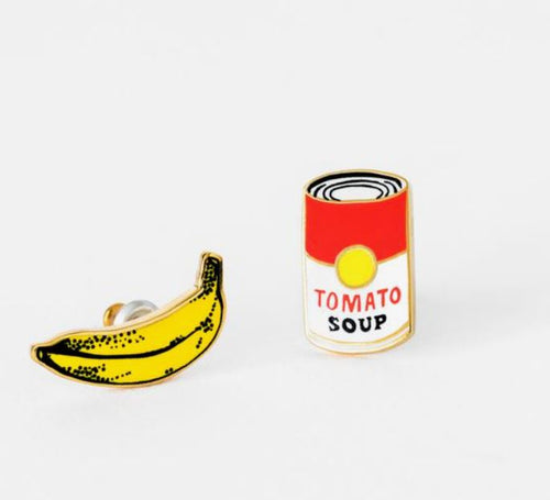 The Pop Art Banana & Soup Earrings by Blue Dog Posters are a quirky pair of cloisonné enamel studs on a white backdrop. One earring features a bright yellow banana with black detailing, while the other is designed as a red and white can of tomato soup with "TOMATO SOUP" written on the front, making them unique and collectible accessories.