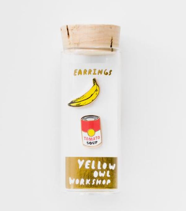 A clear glass vial with a cork stopper contains two cloisonné petite studs from Blue Dog Posters' Pop Art Banana & Soup Earrings. One is shaped like a banana, and the other like a can of tomato soup. The words "EARRINGS" and "YELLOW OWL WORKSHOP" are written on the vial.