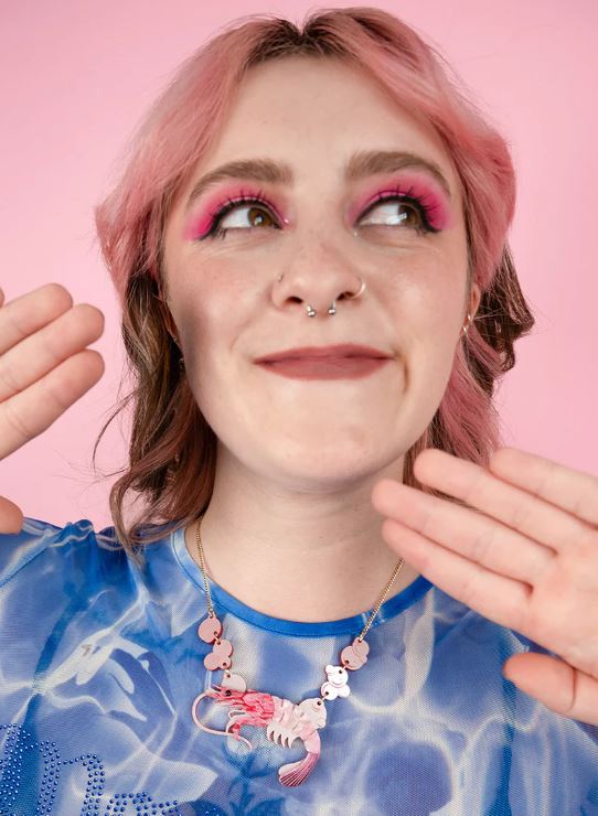 A person with pink hair and vibrant pink eyeshadow is smiling while looking upwards. They have a nose ring and septum piercings. They are wearing a blue patterned shirt and a Pearly Prawn Necklace Pink from Blue Dog Posters, featuring beads and a colorful shrimp pendant. The background is pink.