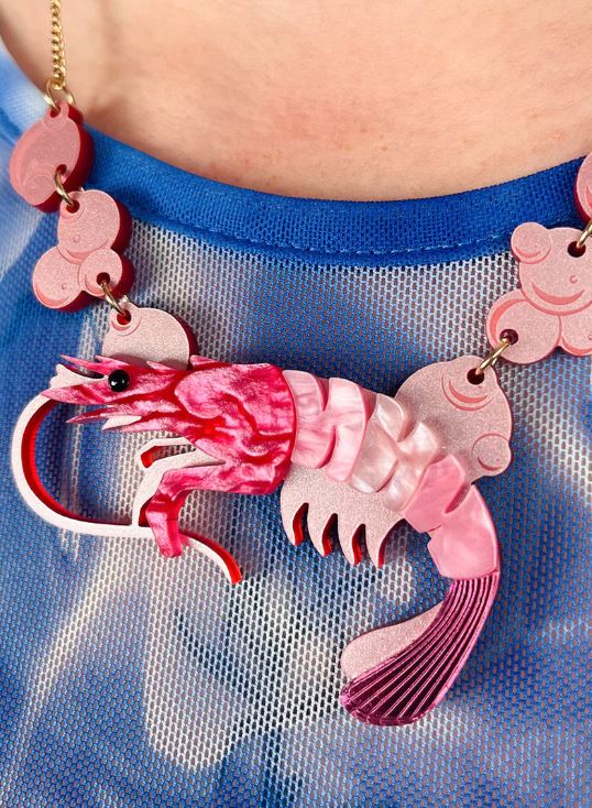 A close-up image shows a person wearing the Pearly Prawn Necklace Pink from Blue Dog Posters, showcasing a unique large pink and red shrimp pendant. The necklace is detailed with intricate designs, including a curled tail and extended antennae, attached to a blue mesh top by an acclaimed UK jewelry designer.