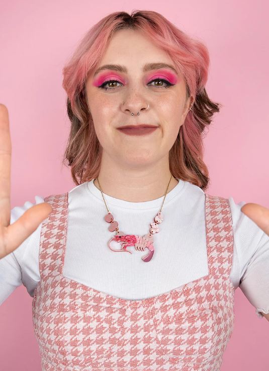 A person with pink-dyed hair and pink eyeshadow is wearing a white top underneath a pink houndstooth dress. They have a Pearly Prawn Necklace Pink from Blue Dog Posters and are smiling while extending their hands forward. The background is light pink.