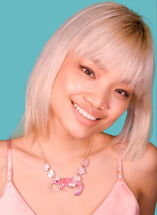 A person with platinum blonde hair and bangs smiles at the camera, against a turquoise background. They are wearing a pink tank top and a unique Pink Pearly Prawn Necklace by Blue Dog Posters, which features a cat-like design from a renowned UK Jewellery Designer.