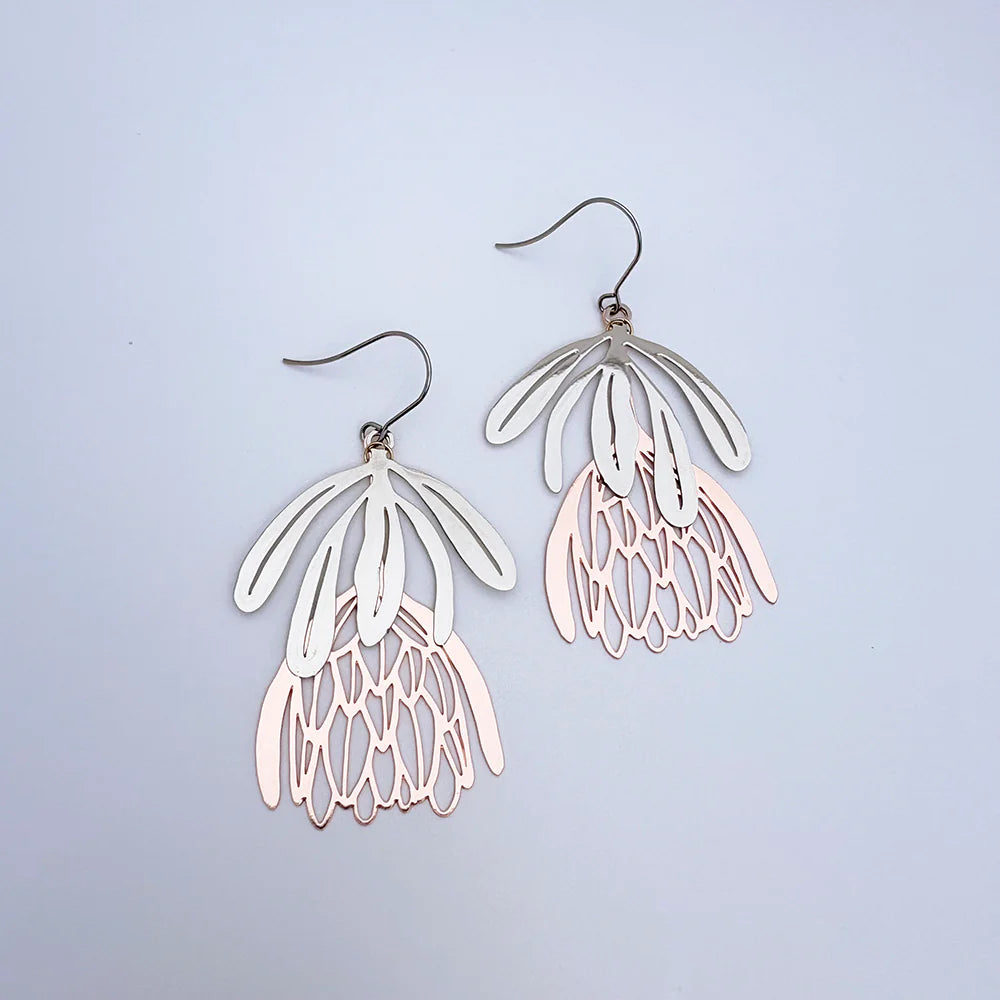 The Protea Flower Silver Rose Gold Denz Earrings by Blue Dog Posters feature layered, petal-like metal shapes in silver and light rose gold tones, forming stylized flowers against a plain light background.