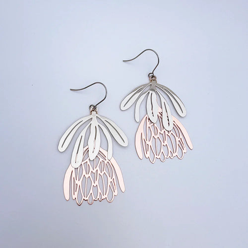 The Protea Flower Silver Rose Gold Denz Earrings by Blue Dog Posters feature layered, petal-like metal shapes in silver and light rose gold tones, forming stylized flowers against a plain light background.