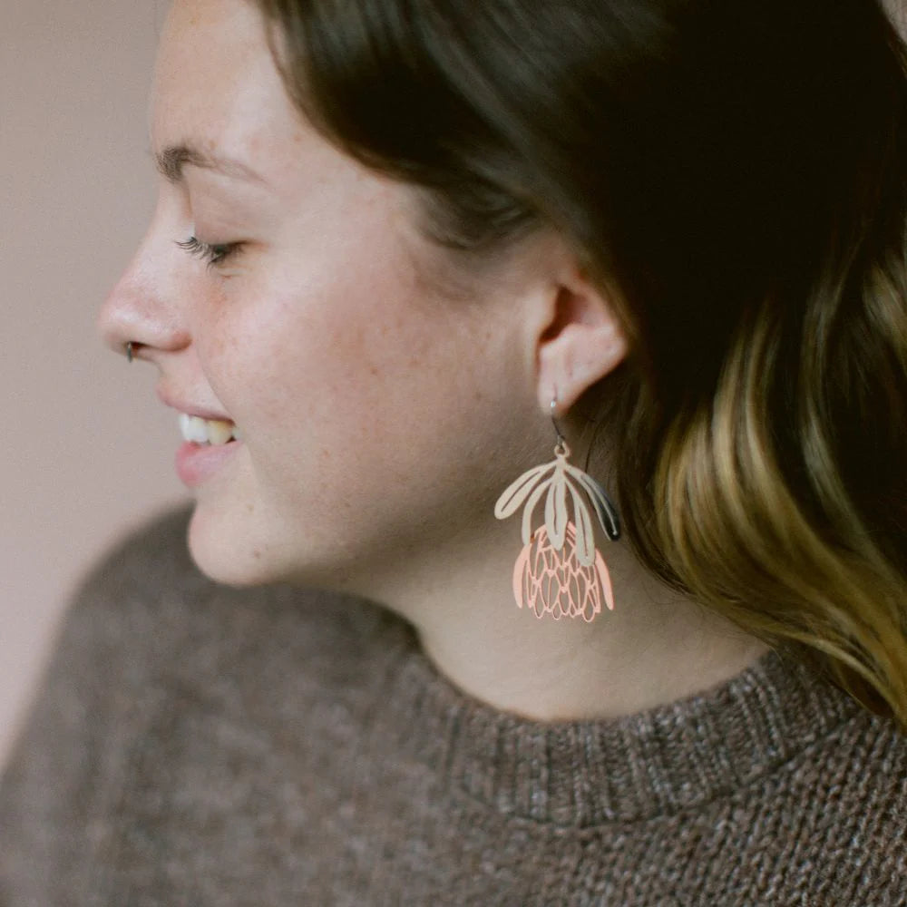 A smiling person with long brown hair and a nose ring wears Blue Dog Posters’ Protea Flower Silver Rose Gold Denz Earrings, paired with a brown knitted sweater. The side-profile photo has a neutral background.