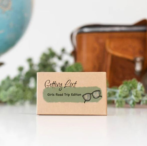 A prominently displayed box labeled "Getting Lost Cards - Girls Road Trip Edition" by Blue Dog Posters sits in focus next to Misdirection Cards. In the background, a globe, a brown leather bag, and some greenery evoke a sense of wanderlust.