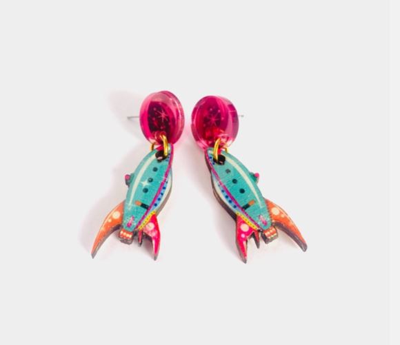 The Little Retro Rocket Earrings by Blue Dog Posters feature turquoise bodies, red-orange fins, and multicolored details on colorful rocket-shaped earrings with bright pink circular studs, set against a white background.