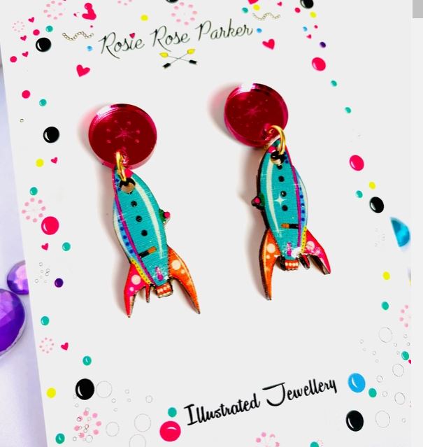 The Little Retro Rocket Earrings by Blue Dog Posters feature vibrant turquoise, orange, and pink rockets hanging from red circular studs. They are displayed on a white card adorned with colorful dots and the text "Rosie Rose Parker Illustrated Jewellery.