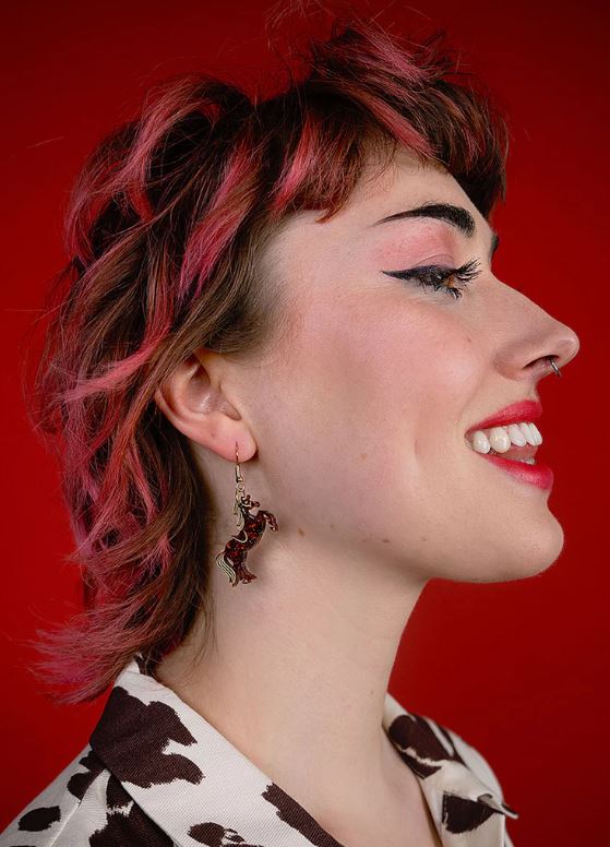 A person with pink-tinted, wavy hair in front of a red background smiles while looking to the side. They wear eyeliner, pink lipstick, and a cow-patterned shirt. Adorned with Blue Dog Posters' Rodeo Horse Earrings and a small nose ring, their whimsical style shines through.