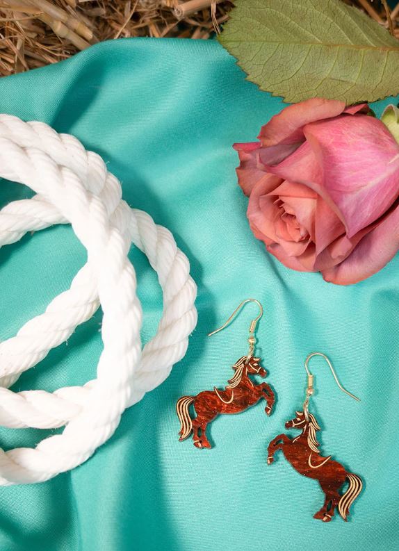 A pair of Rodeo Horse Earrings from Blue Dog Posters, featuring red and gold detailing, lies on a teal fabric next to a coiled white rope and a pink rose. A large green leaf is partially visible in the background, highlighting the intricate lasercut tortoiseshell acrylic design of these stunning accessories.