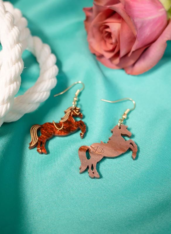A pair of Rodeo Horse Earrings from Blue Dog Posters, crafted with one in copper and the other in wood, are displayed on a turquoise fabric. A pink rose and a coil of white rope lie beside the earrings, evoking the whimsical designs reminiscent of Tatty Divine.