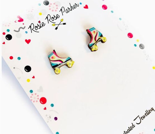 Earrings shaped like colorful roller skates are displayed on a white, illustrated card with a playful, colorful border. The card features the brand name "Blue Dog Posters" and showcases the "80s Roller Boot Retro Stud Earrings.