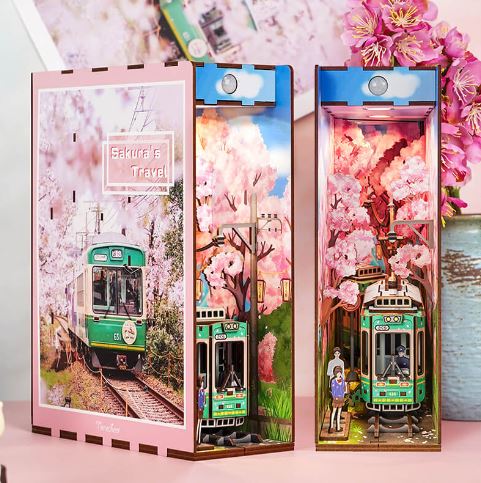 Sakura's Travel Book Nook Tonecheer by Blue Dog Posters features a green tram beneath cherry blossoms with miniature people nearby—a charming Japan-inspired bookshelf insert that brings Kyoto travel vibes to your shelf. The pink sign reads "Sakura's Travel.