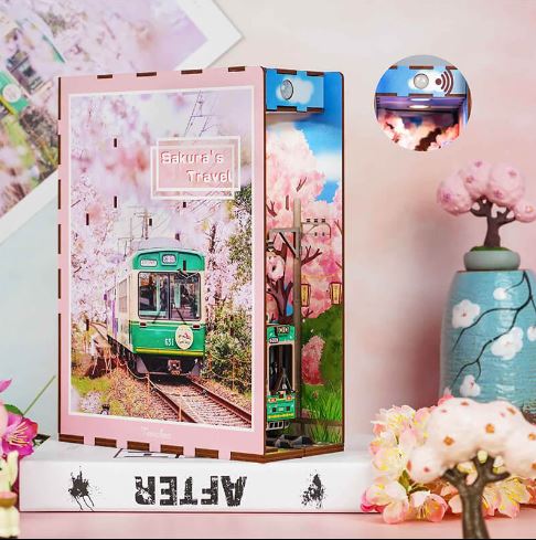 The Sakura’s Travel Book Nook by Blue Dog Posters features a green tram among cherry blossoms, inspired by Kyoto, sitting atop a "FESTER" book with vases and faux flowers, and includes a small light in the top corner.