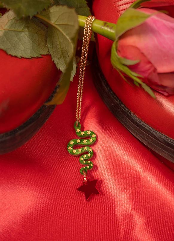 The Lonestar Snake Pendant by Blue Dog Posters, featuring a green snake surrounded by star details, lies gracefully next to a red rose on vibrant satin. In the backdrop, red shoes and lush green leaves offer a captivating contrast.