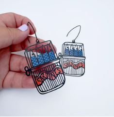 A hand holds two Denz Sardine Tin Earrings by Blue Dog Posters, shaped like sardine cans. The detailed design includes sardines and "SARDINES" in a playful, artistic style against a plain background.