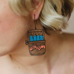 A person with short, blonde hair wears Denz Sardine Tin Earrings by Blue Dog Posters, featuring a novelty sardine can design with blue and silver fish and a "SARDINES" label.
