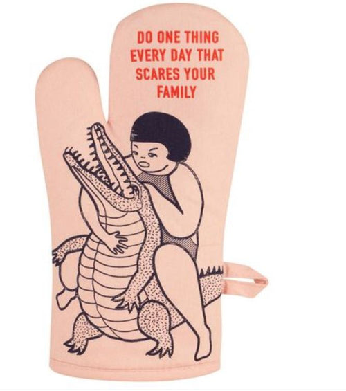 The "Scares Your Family" oven mitt by Blue Dog Posters is a super-insulated pink mitt featuring a black-and-white illustration of a person wrestling an alligator. Bold red text above the image reads, "DO ONE THING EVERY DAY THAT SCARES YOUR FAMILY," and it includes a convenient loop for hanging on the side.