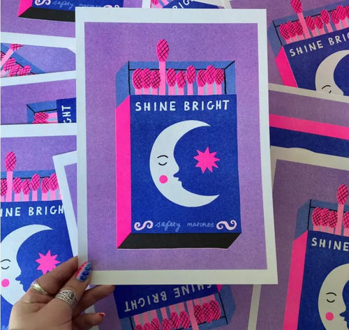 A hand holds an illustrated design of the "Shine Bright A4 Risograph - Amy Hastings" on recycled coffee cup paper, featuring a crescent moon and star. The matchbox is predominantly blue and pink, while several identical Risograph prints from Blue Dog Posters are scattered in the background.