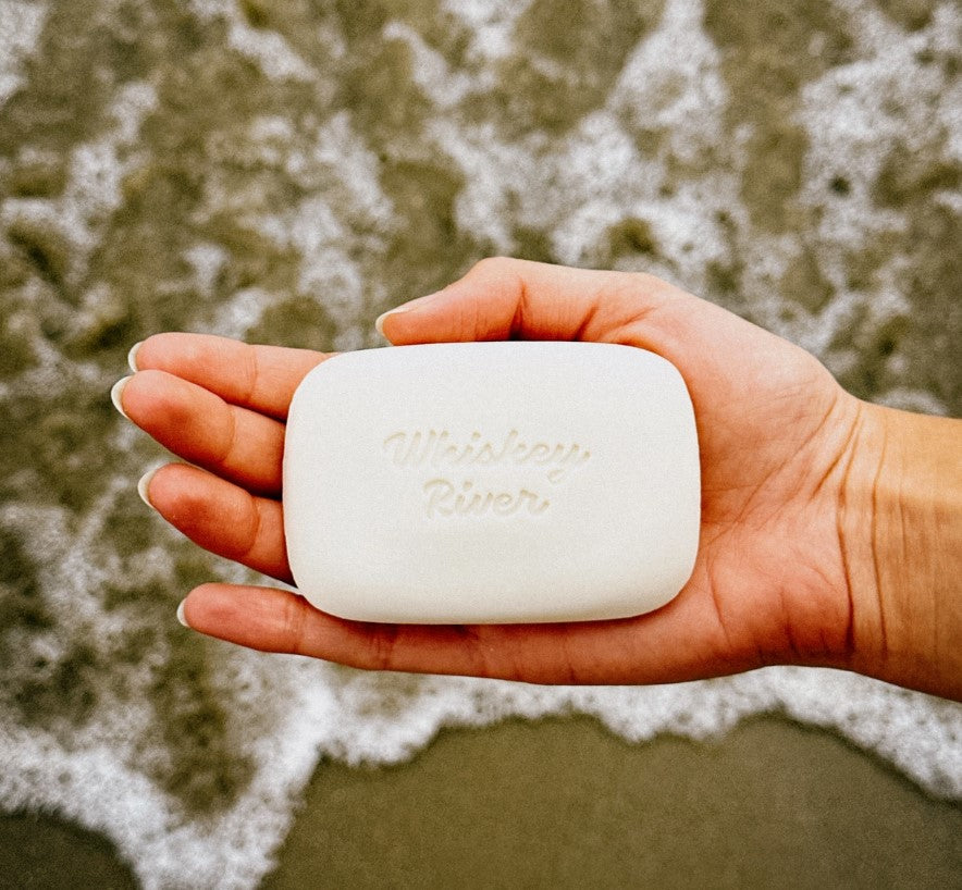 A hand holds a rectangular white soap bar with the embossed text "Cunt Triple Milled" from Blue Dog Posters while standing near the ocean. The background shows waves crashing on the shore, capturing the essence of their vintage-style box packaging.