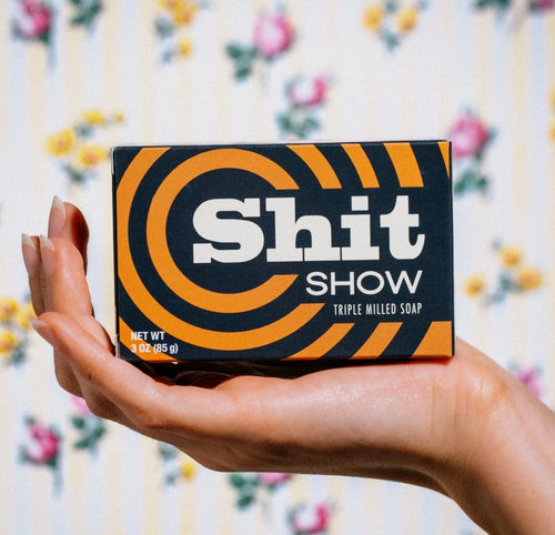 A hand holds a Vermont-made Shit Show Triple Milled Boxed Bar Soap from Blue Dog Posters, adorned with bold text "Shit Show." The vintage-style box is decorated with a black and orange swirl design. Against floral wallpaper featuring delicate pink and yellow flowers, this 3 oz (85 g) soap promises top-notch personal hygiene.