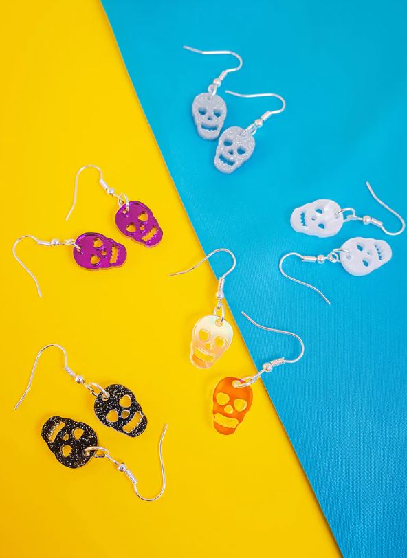 A collection of Blue Dog Posters' Skull Charm Earrings Purple Mirror displayed on a bright, contrasting background. These whimsical designs come in various colors including purple mirror, pink, black, yellow, white, and glittery styles with silver hooks. The vibrant backdrop is split into yellow and blue sections.