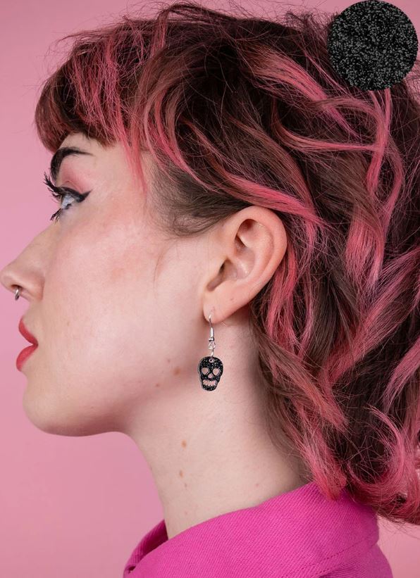 A person with pink hair and a nose piercing is wearing Blue Dog Posters' Skull Charm Earrings in Black Glitter. They are dressed in a pink shirt, posed against a pink background, looking to the right.
