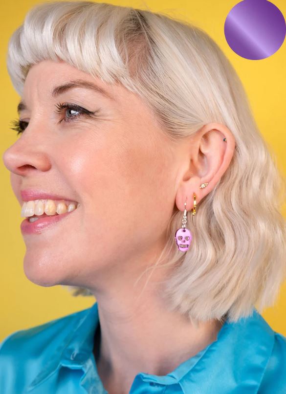 A person with short, wavy blonde hair smiles while looking to the left. They are wearing a blue shirt and a purple Skull Charm Earring in their left ear from Blue Dog Posters. The background is a solid yellow color, and there is a small purple circle in the upper right corner.