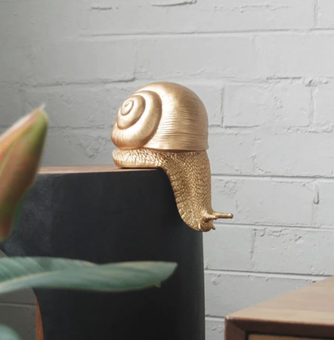 A friendly Shelf Snail - Gold sculpture from Blue Dog Posters, crafted as a resin ornament, is perched on the edge of a dark-colored rectangular shelf against a light gray brick wall. A green plant with a budding flower adds charm to the home décor in the foreground on the left side of the image.