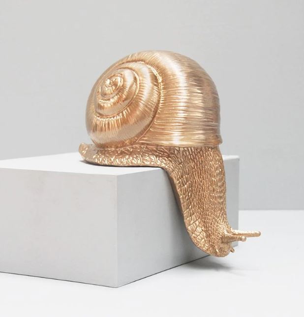 The Shelf Snail - Gold by Blue Dog Posters, perfect for enhancing home décor, is elegantly placed on a white, rectangular pedestal against a simple gray backdrop. This charming figurine boasts intricate textures on its shell and body, showcasing the gleaming and reflective qualities of its metallic gold finish.