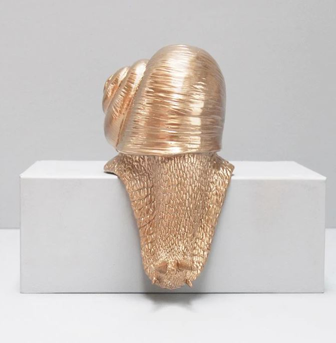 The "Shelf Snail - Gold" by Blue Dog Posters is a metallic sculpture featuring a friendly snail with an intricately detailed, textured body and shell, set atop a plain white rectangular pedestal. This home décor piece, made of shiny gold material, stands out brilliantly against a neutral gray background.
