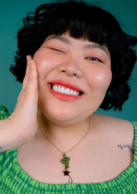 A smiling person with short black hair, dressed in a green top, poses against a teal background. With one hand resting on their cheek, they elegantly display the Lonestar Snake Pendant by Blue Dog Posters, while a tattoo is visible on their shoulder.