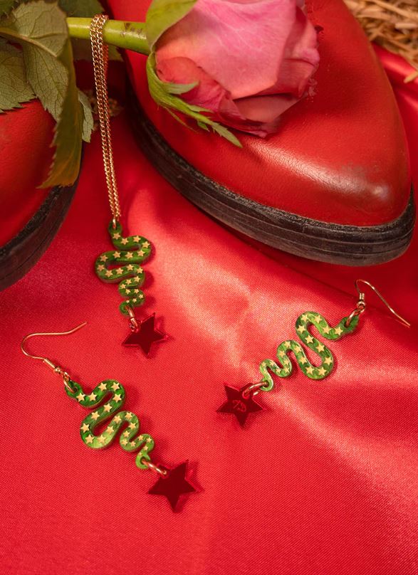 The Lonestar Snake Pendant earrings from Blue Dog Posters, featuring green snake designs with red star accents crafted from laser-cut acrylic, are elegantly showcased on luxurious red satin fabric beside a single pink rose and vibrant red shoes.