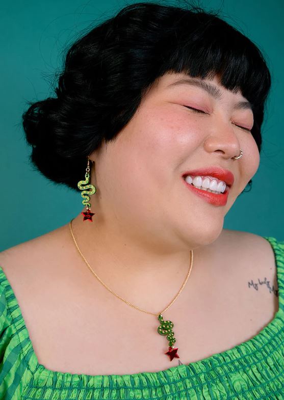 A person with dark hair smiles joyfully against a teal background, wearing a green off-shoulder top. Their nose ring complements the laser-cut acrylic earrings and the Lonestar Snake Pendant by Blue Dog Posters, featuring a green snake and red star design, elegantly hanging from a gold-plated chain.