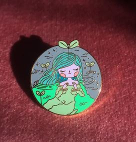 A round enamel pin featuring a cartoon character with green hair and a plant sprout on their head, sitting on a rock amidst water. The Soft n Sweet Enamel Pin by Blue Dog Posters showcases a peaceful character surrounded by small sprouts and stars, all enhanced with a shiny gold metal finish.