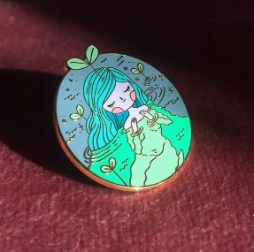 The Soft n Sweet Enamel Pin by Blue Dog Posters portrays a girl with blue-green hair, holding two rings. Adorned with a small plant sprout on her head, she is surrounded by green butterflies, and her calm expression stands out against a gradient blue and green background. Finished in shiny gold metal and designed in Melbourne by Hannakin, this exquisite pin captures an enchanting scene.