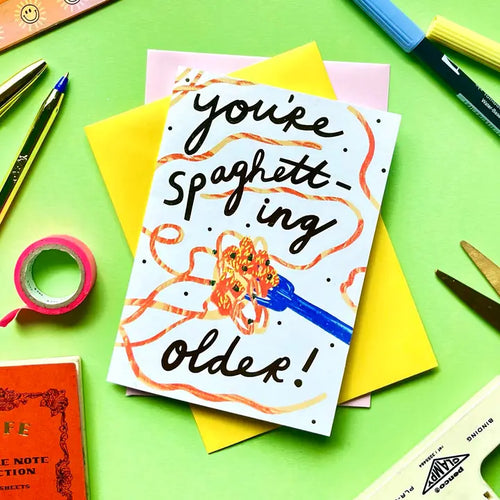 The You're Spaghetting Older Greeting Card by Blue Dog Posters features a spaghetti-on-fork illustration and the punny message "You're Spaghetti-ing Older!" on a green surface, surrounded by pens, scissors, tape, and envelopes.