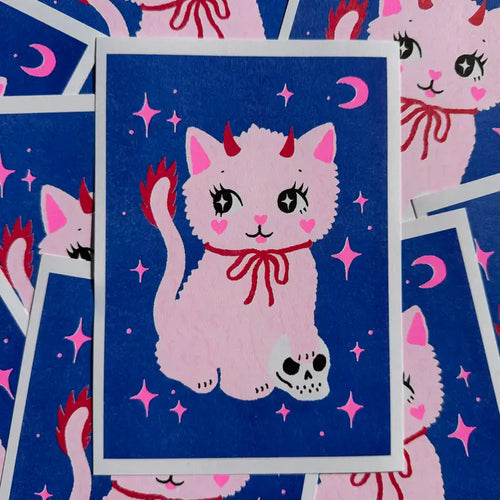 Cute illustration of a fluffy pink cat with red devil horns, tail, red bow, and skull by its paw on a blue background with pink stars and moons. "Spooky Cat A5 Risograph - Amy Hastings" by Blue Dog Posters, printed on recycled coffee cup paper.