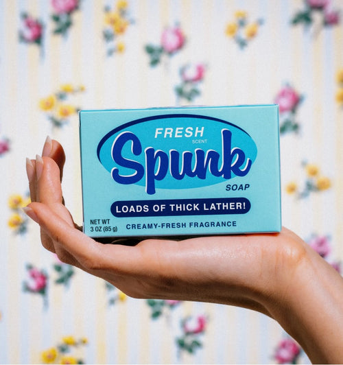 A hand holds a vintage-style blue box of Fresh Spunk Triple Milled Boxed Bar Soap by Blue Dog Posters against a floral wallpaper background. The packaging reads, "Fresh scent Spunk soap. Loads of thick lather! Net wt 3 oz (85g). Creamy-fresh fragrance." It complements the colorful flowers in the background.